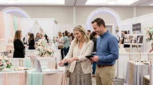 Couple souriant explorant les stands d'un salon du mariage. La femme pointe un dépliant, l'homme tient un carnet. Décors fleuris et gâteaux.